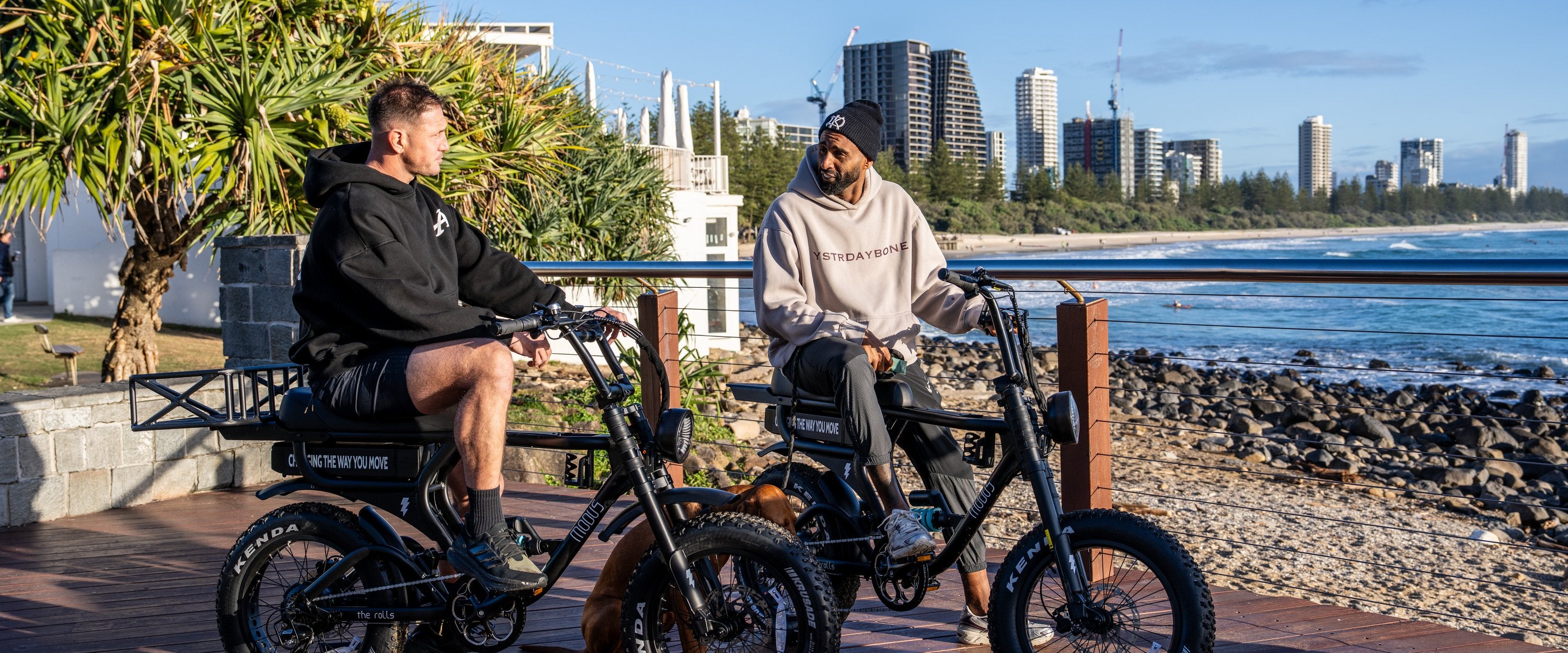 Two riders sitting on their Rolls e-bikes on the Burleigh Heads boardwalk, Gold Coast skyline across the water