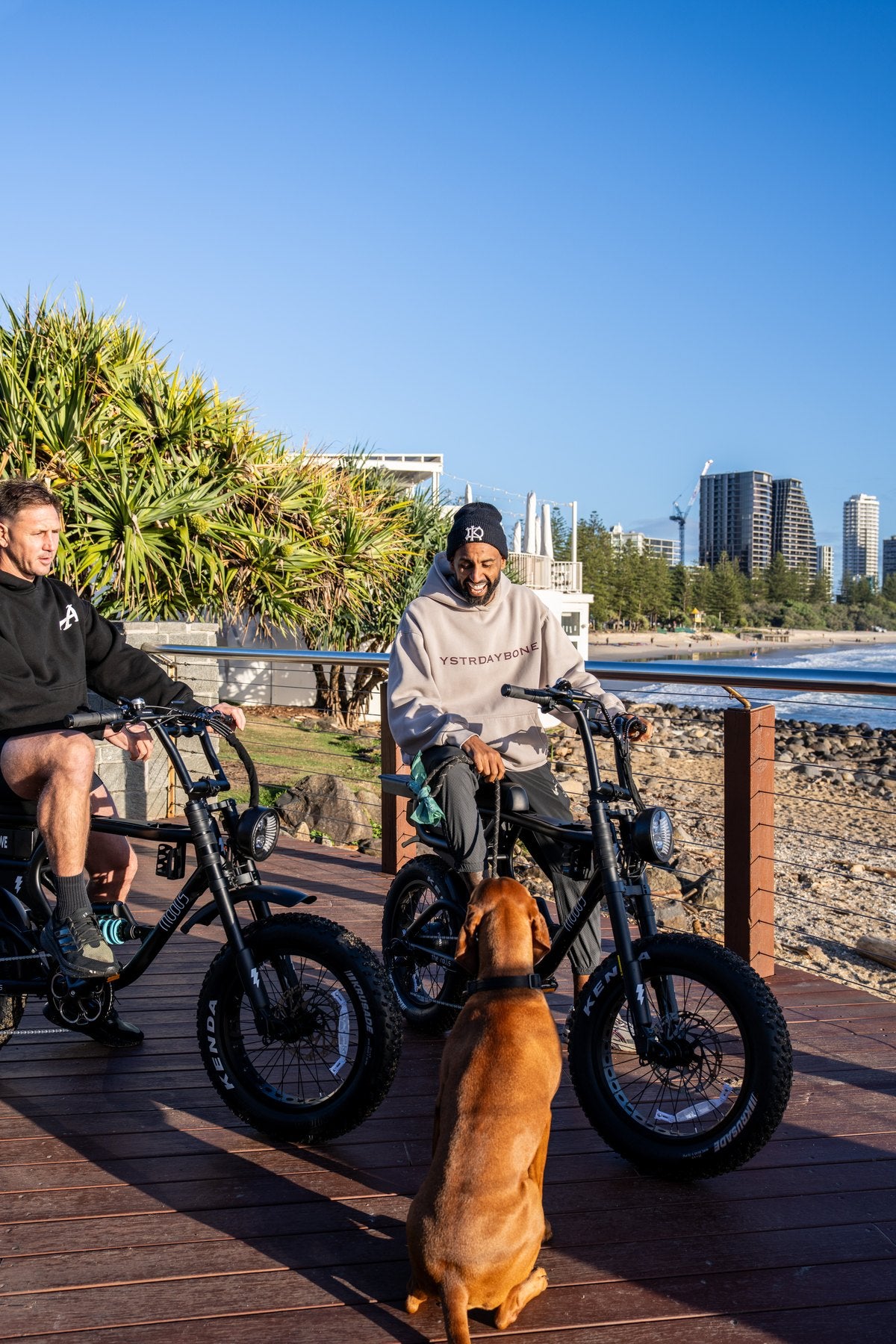 Two riders on Rolls e-bikes with a dog on the Burleigh boardwalk